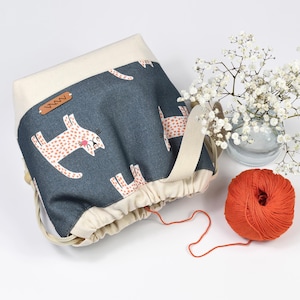 Può includere: Una borsa blu denim con coulisse, con la parte superiore e il cinturino color crema. La borsa presenta un motivo ripetuto di gatti dei cartoni animati con macchie rosa. Una palla di filo arancione e un piccolo vaso di fiori bianchi sono accanto alla borsa.