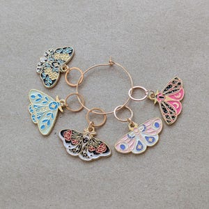 Op de afbeelding: Vijf gouden metalen vlindercharms met kleurrijke emaildetails hangen aan een gouden metalen ring. De vlinders hebben verschillende patronen en kleuren, waaronder blauw, roze, zwart en wit.