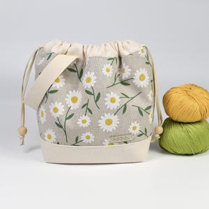 Könnte beinhalten: Ein beigefarbener Beutel mit Kordelzug und einem Gänseblümchen-Blumenmuster. Die Tasche hat einen cremefarbenen Boden, einen Kordelzugverschluss und einen passenden Griff. Zwei Wollknäuel, eines gelb und eines grün, liegen neben der Tasche.
