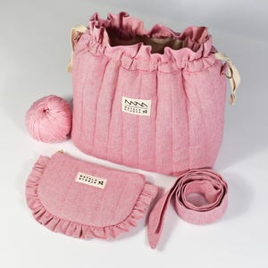 Op de afbeelding: Een roze gewatteerde tas met trekkoord, een geplooide bovenkant en crèmekleurige linten, een bijpassend halfrond tasje met rits en een geplooide rand, een roze bol wol en een lange roze riem. Elk item is voorzien van een label met Natala Studio.