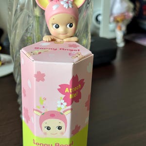 May include: A Sonny Angel figurine with a pink deer-themed hat and a matching box. The box is pink with cherry blossom illustrations and the text "Attach Me!". The figurine is a collectible item.