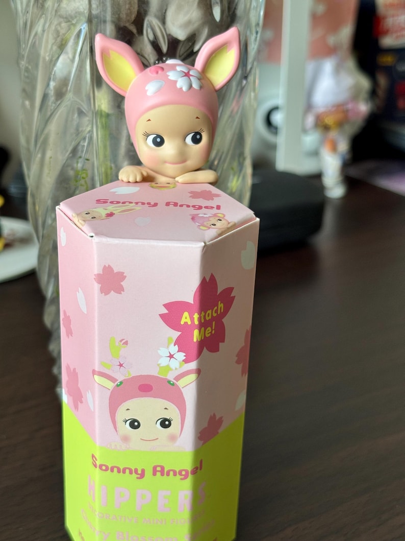 May include: A Sonny Angel figurine with a pink deer-themed hat and a matching box. The box is pink with cherry blossom illustrations and the text "Attach Me!". The figurine is a collectible item.