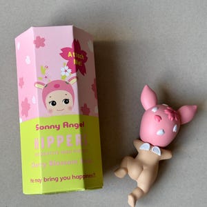 May include: A Sonny Angel Cherry Blossom series figurine and its packaging. The figurine is a pink bunny with white accents and small wings. The box is pink and green with the words "Sonny Angel" and "HIPPER" printed on it.