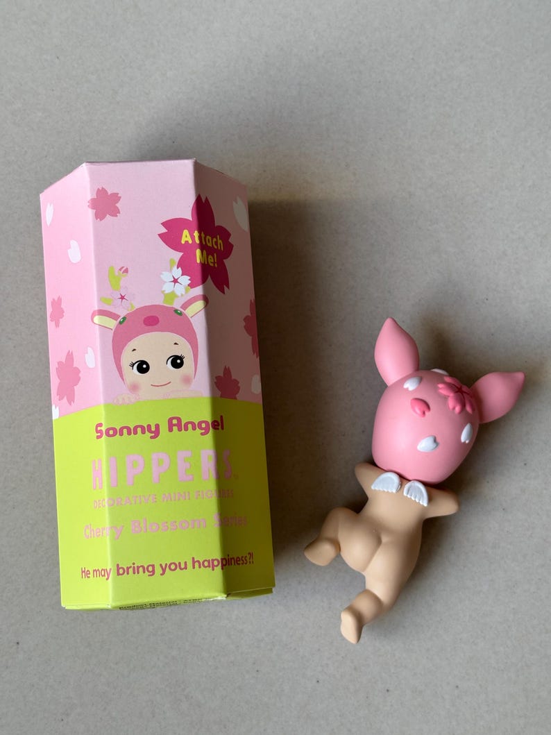 May include: A Sonny Angel Cherry Blossom series figurine and its packaging. The figurine is a pink bunny with white accents and small wings. The box is pink and green with the words "Sonny Angel" and "HIPPER" printed on it.
