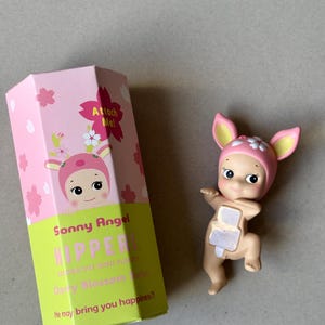 May include: A Sonny Angel Hipper figurine with a pink and white floral hat and matching box. The box reads "Attach Me!" and "He may bring you happiness?" The figurine is a light tan color.