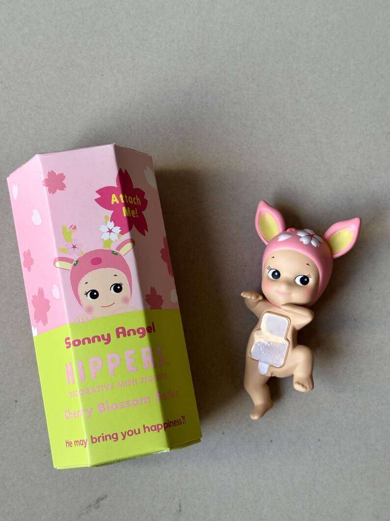 May include: A Sonny Angel Hipper figurine with a pink and white floral hat and matching box. The box reads "Attach Me!" and "He may bring you happiness?" The figurine is a light tan color.