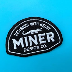 Puede incluir: Parche bordado en blanco y negro con el texto "DESIGNED WITH HEART" y "MINER DESIGN CO." y un gráfico de cocodrilo blanco.