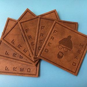 Könnte beinhalten: Ein Stapel brauner Lederpatches mit dem Text "FRED HEAD" und einer Cartoon-Illustration einer Person mit Hut, Brille und Bart.