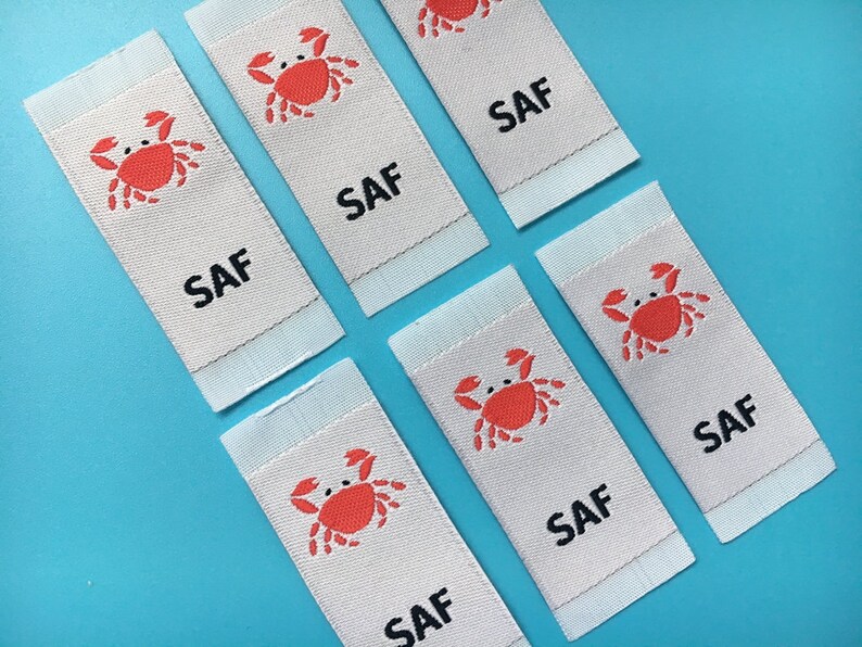 May include: Six white fabric labels with a red crab design and the word "SAF" printed in black.