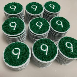 Peut inclure: Une collection d'écussons circulaires disposés en huit piles. Chaque écusson présente une surface vert foncé texturée avec un chiffre neuf brodé en blanc au centre et une bordure en feutre blanc autour du bord de la forme circulaire.