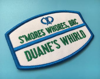 May include: Embroidered patch with a white background, blue border, and blue and green text. The text reads "S'MORES WHORES, BRC" and "DUANE'S WHIRLD". The patch has a unique shape and is likely for a jacket or bag.