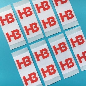 May include: A close-up of ten white woven labels with the letters "HB" printed in red.