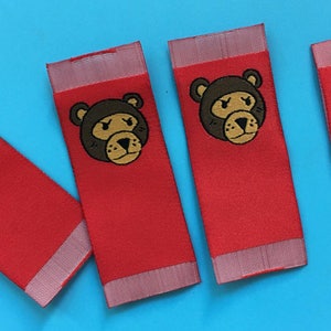 May include: Five red fabric labels with white trim. Three labels feature a brown bear with a gold face and black eyes. Two labels feature a white "W" with a dripping paint effect.