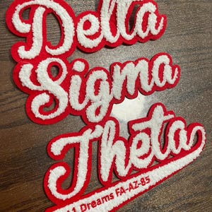 May include: A red and white chenille patch featuring the text Delta Sigma Theta in a cursive script font. The patch includes a white tail section with the red text 11 Dreams FA-AZ-85 embroidered on it, with a red border outlining the entire design.
