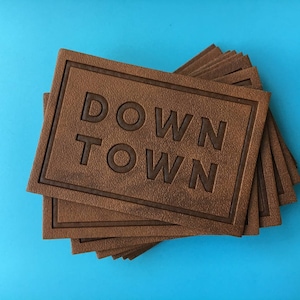 Könnte beinhalten: Ein Stapel brauner rechteckiger Lederaufnäher mit dem geprägten Schriftzug "DOWN TOWN" in der Mitte. Die Aufnäher haben eine strukturierte Oberfläche und einen dunkelbraunen Rand. Der Hintergrund ist einfarbig hellblau.