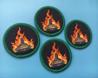 May include: Four green and black embroidered patches featuring a UFO flying through flames. The UFO is dark grey and the flames are orange and yellow.