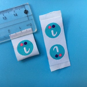 May include: Two white fabric labels with a turquoise circle containing the letter "I" and three small dots in pink, purple, and black.
