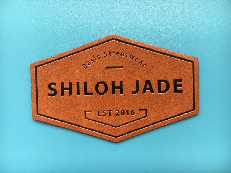 May include: A brown leather patch with black stitching in the shape of a hexagon. The text "Basic Streetwear" is at the top, followed by "SHILOH JADE" in a larger font, and "EST 2016" at the bottom.