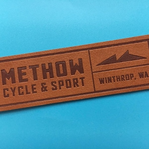 Könnte beinhalten: Braunes Lederetikett mit dem Text "Methow Cycle & Sport" und "Winthrop, WA" in weißen Buchstaben. Das Etikett zeigt eine stilisierte Grafik einer Bergkette.