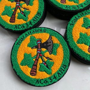 Pode incluir: Patches bordados circulares com um centro amarelo, um machado tomahawk preto e folhas verdes. A borda é verde com o texto preto TOMAHAWKS e ACo. 34 AHB. Os patches têm uma borda preta e uma superfície texturizada em relevo.