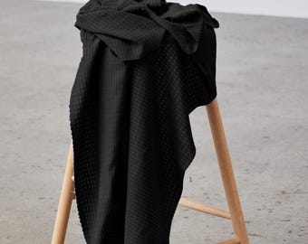 May include: A black waffle-weave fabric draped over a light wooden stool. The fabric is textured with a grid pattern and hangs down the side of the stool. The stool has three legs and a simple design.