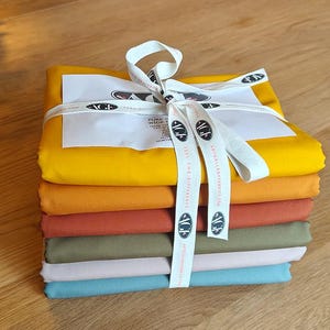 Puede incluir: Un conjunto de siete muestras de tela en varios tonos de amarillo, naranja, marrón, verde, rosa y azul. La tela está atada con una cinta blanca con el texto "PURE WIDE WIDTH FABRIC" y "FEEL THE DIFFERENCE" impreso en ella.