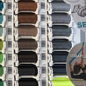 Könnte beinhalten: Nahaufnahme einer Wandanzeige mit Garnrollen in verschiedenen Farben. Die Spulen sind in Reihen und Spalten angeordnet, und jede Spule hat ein Etikett mit einer Nummer und einem Barcode. Der Text "FANTASTIC ELASTIC" und "FANTASTISCH ELASTISCH" ist auf einem weißen Kreis sichtbar.