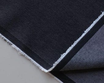 May include: Close-up of a dark navy blue denim fabric with a textured surface. The fabric has a raw, unfinished edge with white threads. A folded section reveals a darker, almost black, stripe.