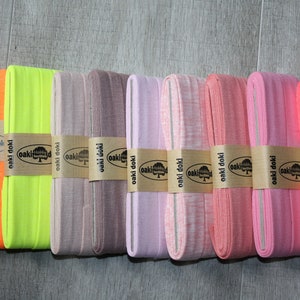 Op de afbeelding: Acht rollen elastische stof in verschillende kleuren met de tekst "oaki doki" gedrukt op het etiket. De stof is verkrijgbaar in oranje, geel, lichtbruin, lichtpaars, roze, lichtroze en rood.