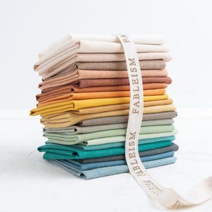Op de afbeelding: Een stapel gevouwen stoffen vierkanten in een kleurverloop van crème en beige tot geel, groen en blauw. Een wit lint met de herhalende tekst FABLEISM is verticaal over het midden van de stoffen stapel gedrapeerd.