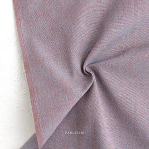 Könnte beinhalten: Ein Stück Webstoff in einer Farbpalette aus gedämpftem Mauve und staubigem Blau. Das Textil weist eine sichtbare Kreuzgewebestruktur mit einer weichen, matten Oberfläche auf. Das Wort FABLEISM ist in kleinen weißen Buchstaben unten mittig gedruckt.