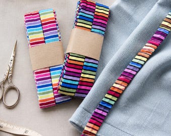May include: Two folded bundles of multi-colored striped fabric trim wrapped in brown paper bands, a single strip of the same striped trim laid across light blue fabric, a wooden spool of blue thread, metal scissors, and a measuring tape.