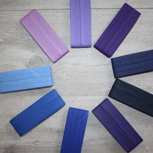 Könnte beinhalten: Acht unterschiedlich farbige Stoffstreifen, die auf einer Holzoberfläche in einem Kreis angeordnet sind. Die Farben sind helllila, lila, dunkellila, dunkelblau, blau, hellblau, dunkelblau und ein dunklerer Lila-Ton.