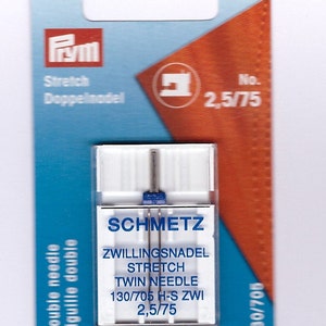 May include: A packet of Schmetz twin needles for stretch fabrics. The white plastic container holds two needles with blue accents. The packaging includes the text "Stretch Twin Needle" and "2.5/75". The package is blue and orange.