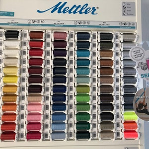 Puede incluir: Una muestra de 72 carretes de hilo de varios colores, etiquetados como "SERAFLEX" y "72 Farben / 72 colours". Los carretes están dispuestos en filas sobre un fondo blanco. El nombre de la marca "Mettler" es visible en la parte superior de la muestra.