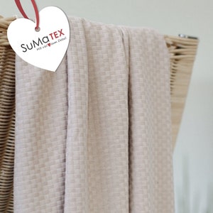 Może przedstawiać: Beżowy materiał wafelkowy o subtelnej fakturze. Materiał jest udrapowany na wiklinowym koszu. Etykieta na materiale głosi "SUMATEX Mit viel zum Detail".