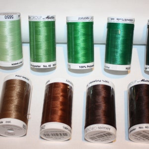 May include: A collection of ten spools of thread in various shades of green and brown. The spools are labeled with the brand name "Amann Group" and "Metler" and the thread type "Polyester".