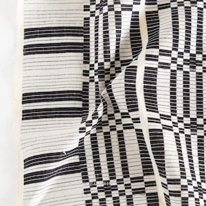 May include: Black and white patterned fabric with a geometric design. The fabric features a mix of horizontal stripes and a checkerboard pattern, creating a visually dynamic textile. The material appears to be woven, with a soft, textured appearance.