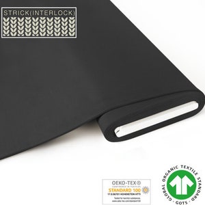Può includere: Rotolo di tessuto nero con il testo "STRICK(INTERLOCK)" e un motivo bianco e grigio. Il tessuto è certificato Oeko-Tex Standard 100 e Global Organic Textile Standard (GOTS).