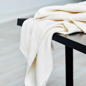 May include: Off-white fabric draped over a black bench. The fabric has a slightly textured appearance and a raw edge. The bench has a minimalist design with a rectangular top and black legs. The scene is lit with natural light.