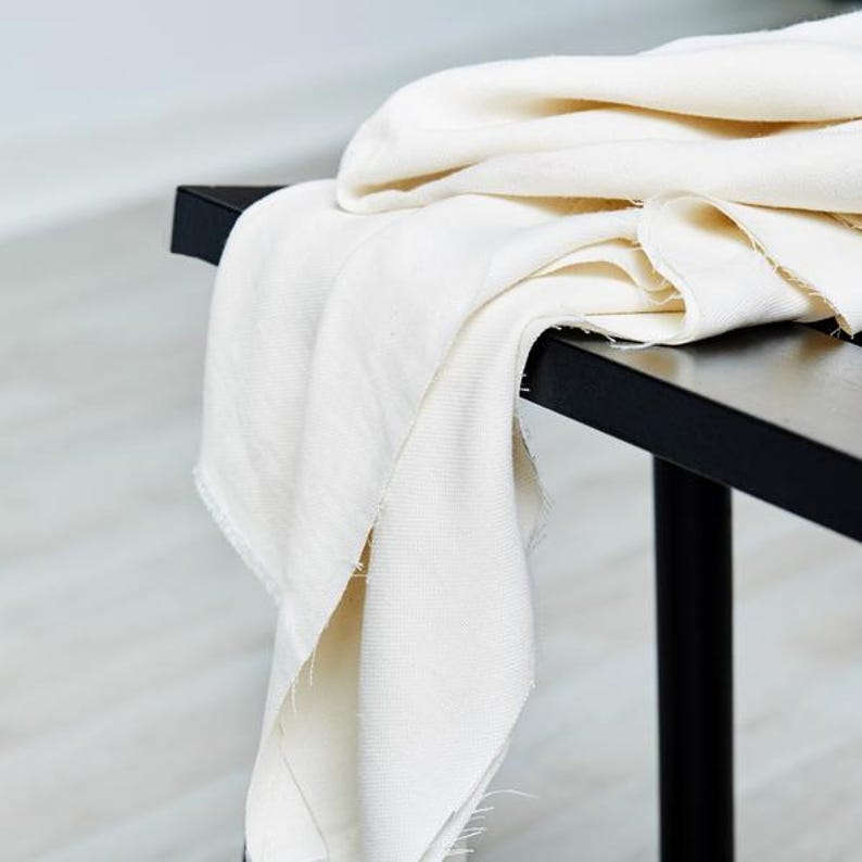May include: Off-white fabric draped over a black bench. The fabric has a slightly textured appearance and a raw edge. The bench has a minimalist design with a rectangular top and black legs. The scene is lit with natural light.