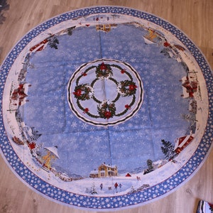 Può includere: Una tovaglia natalizia rotonda, azzurra, con una scena di villaggio invernale con case, alberi e persone. Il bordo presenta un motivo a fiocco di neve. Il centro ha un disegno a ghirlanda. La tovaglia è in tessuto.