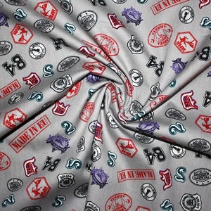Könnte beinhalten: Grauer Stoff mit einem sich wiederholenden Muster aus roten, schwarzen und blauen Stempeln. Die Stempel zeigen verschiedene Designs, darunter Zahlen, Wörter und Symbole. Der Stoff ist in der EU hergestellt.