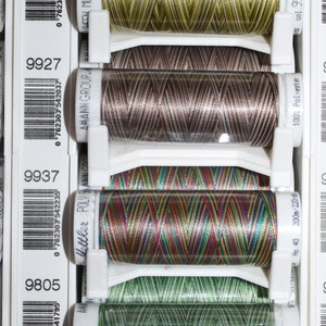 Puede incluir: Primer plano de seis carretes de hilo de colores, cada uno con una etiqueta blanca con un código de barras y un número. El hilo está enrollado alrededor de un carrete de plástico transparente y está dispuesto verticalmente en un recipiente de plástico blanco. Los números en las etiquetas son 9981, 9976, 9927, 9937, 9805 y 9971.