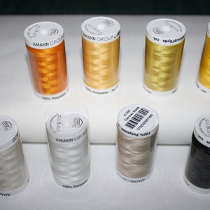 Könnte beinhalten: Acht zylindrische Spulen aus 100 % Polyester-Stickgarn von Amann Group Poly Sheen, angeordnet in zwei Reihen. Die Farben umfassen Orange, Gold, Gelb, Hellbeige, Weiß, Beige und Schwarz, jeweils mit einem weißen Etikett mit Text.