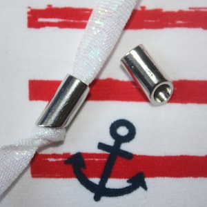 Könnte beinhalten: Zwei silberne Metall-Kordelenden für eine weiße und silberne Kordel. Die Kordelenden befinden sich auf einem weiß-rot gestreiften Stoff mit zwei dunkelblauen Ankern.