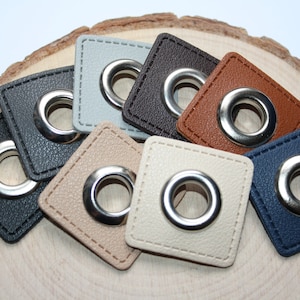Könnte beinhalten: Sortiment von acht quadratischen Lederösen in verschiedenen Farben, darunter Schwarz, Braun, Beige, Blau und Grau. Jede Öse hat einen silbernen Metallring.
