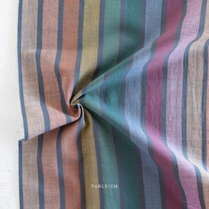 May include: A woven fabric featuring a repeating vertical striped pattern in shades of muted orange, mustard yellow, teal, blue, and deep magenta, separated by thin dark charcoal lines. The word FABLEISM is printed in small white text near the centre.
