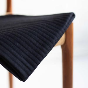 Può includere: Un cuscino per seduta rettangolare nero con trapuntatura a canali orizzontali. Il cuscino è posizionato su una struttura di sedia in legno che presenta un materiale intrecciato di colore chiaro attorno alla parte superiore delle gambe.