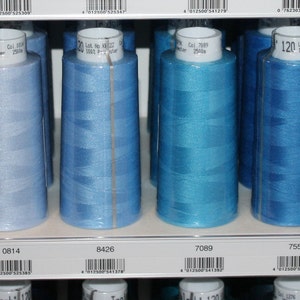 Może przedstawiać: Zbliżenie na dziesięć szpulek nici w różnych odcieniach niebieskiego. Szpulki są ułożone w rzędzie na półce. Szpulki są oznaczone numerami i tekstem "120 m x 100".